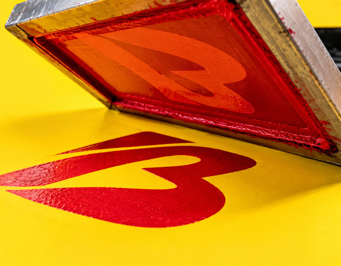 A screenprint screen lifting to reveal a freshly printed red mark on yellow stock.